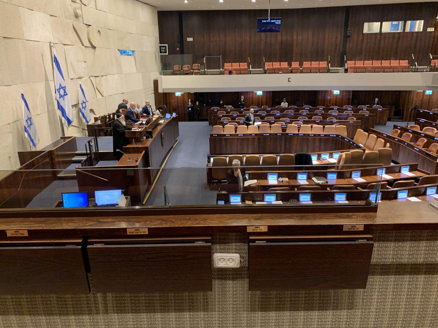 Interior view of Jewish Knesset meeting space with rows of chairs facing a limestone wall with sculptural relief