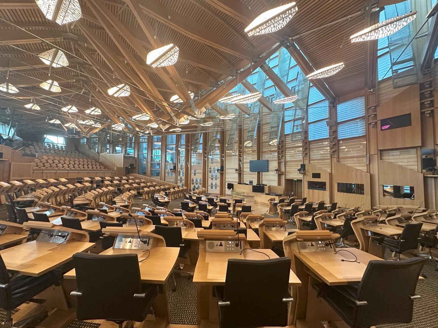 Modern glass and steel meeting space with light wood desks facing a central point, overhung by modern ovular light fixtures in a high wooden slatted ceiling.