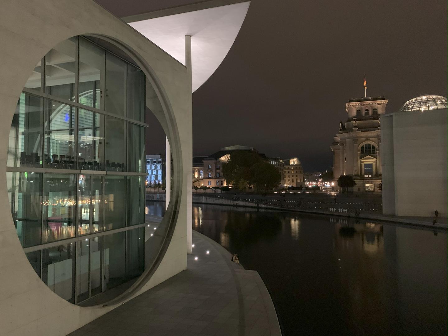 Exterior night view of modern building with multi-story circular window, girded by the curve of a river and standing across from older multi-story buildings.