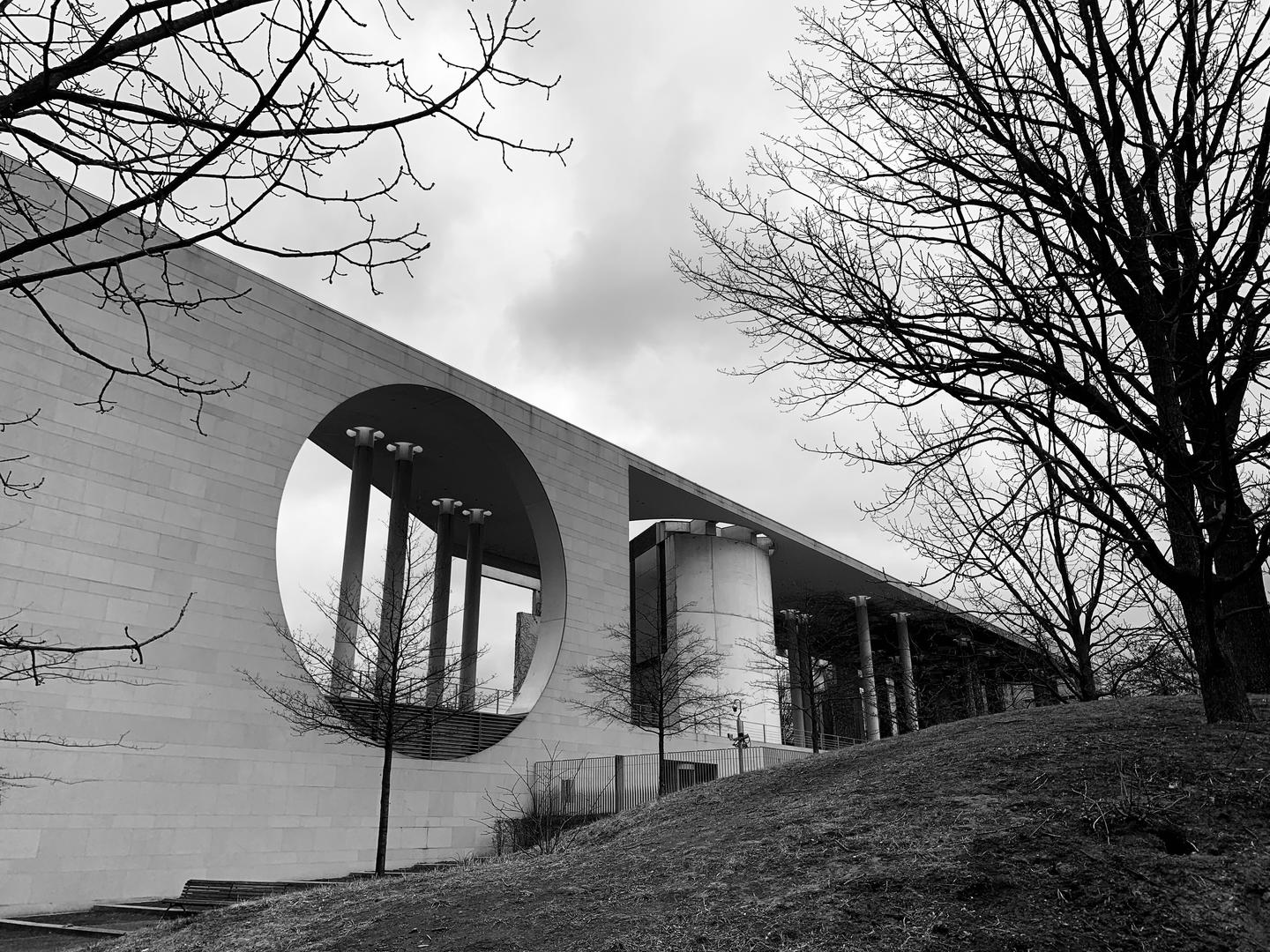 Black and white photo of German Chancellery buidling with a large circular window opening and standing on a hill surrounded by trees.