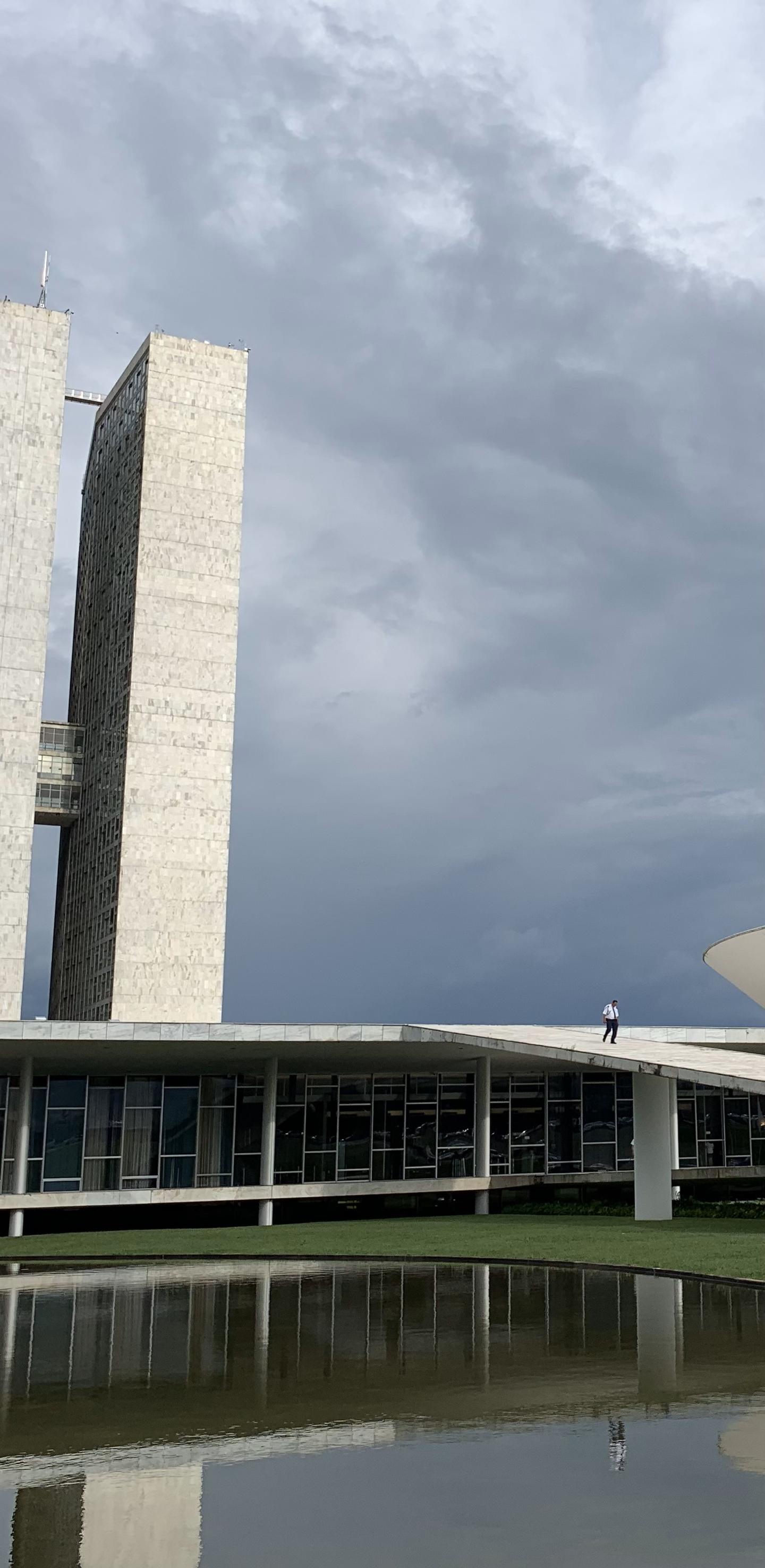 Offset exterior view of the two towers of Brasil's Congresso Nacionale reflected in a body of water under a turbulent cloudy sky.