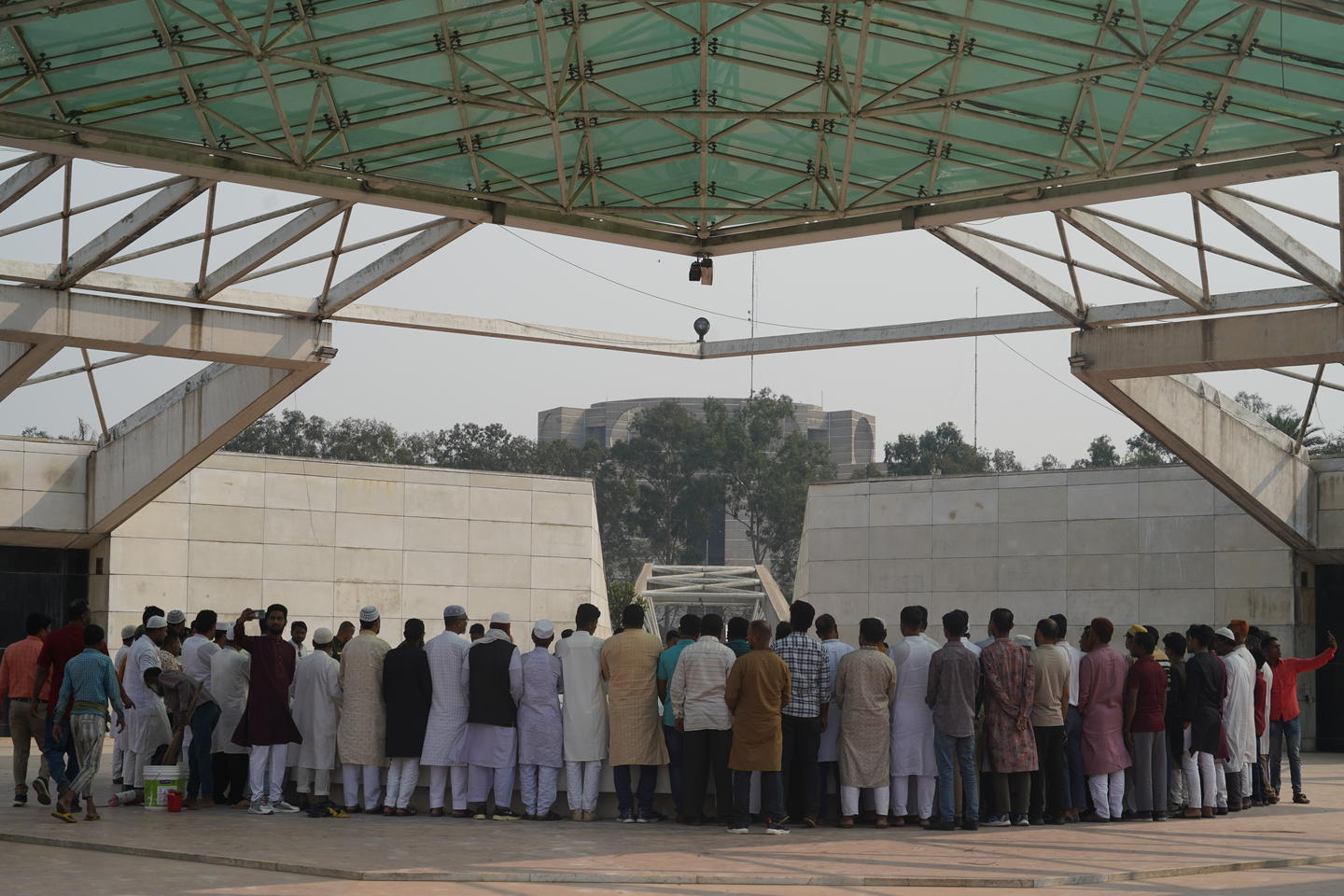 A close-up back view of a crowd of mostly men standing underneath a steel and green glass pavillion at the Mausoleum of Ziaur Rahman.
