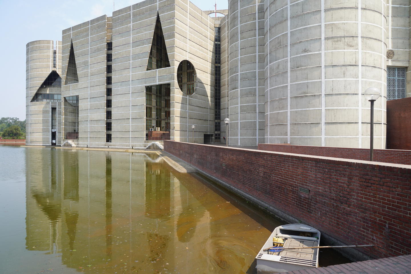 Exterior view of the columnar and triangular-windowed National Assembly Building in Dhaka reflected in a greenish water moat.