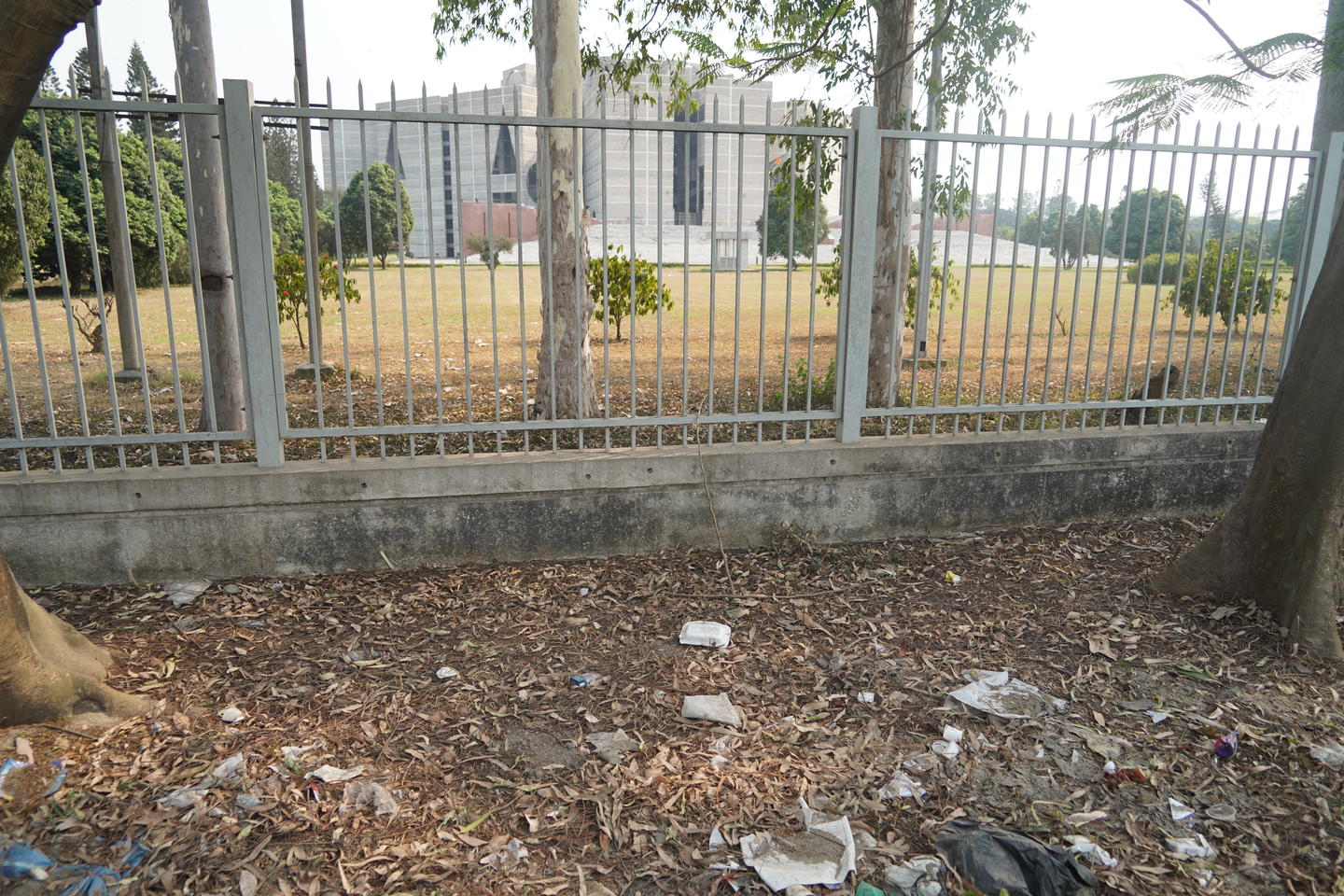 Exterior of National Assembly Building in Dhaka with manicured grounds behind steel and concrete fencing, with litter and leaves strewn in the forefront.