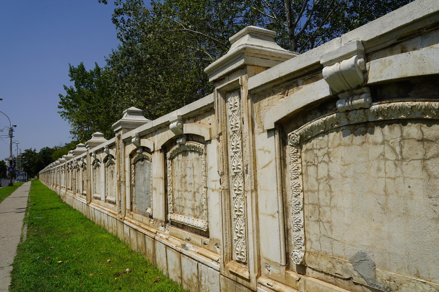 Side view of a white stone carved wall fronted by a strip of grass and a sidewalk, and enclosing tall trees under a blue sky.
