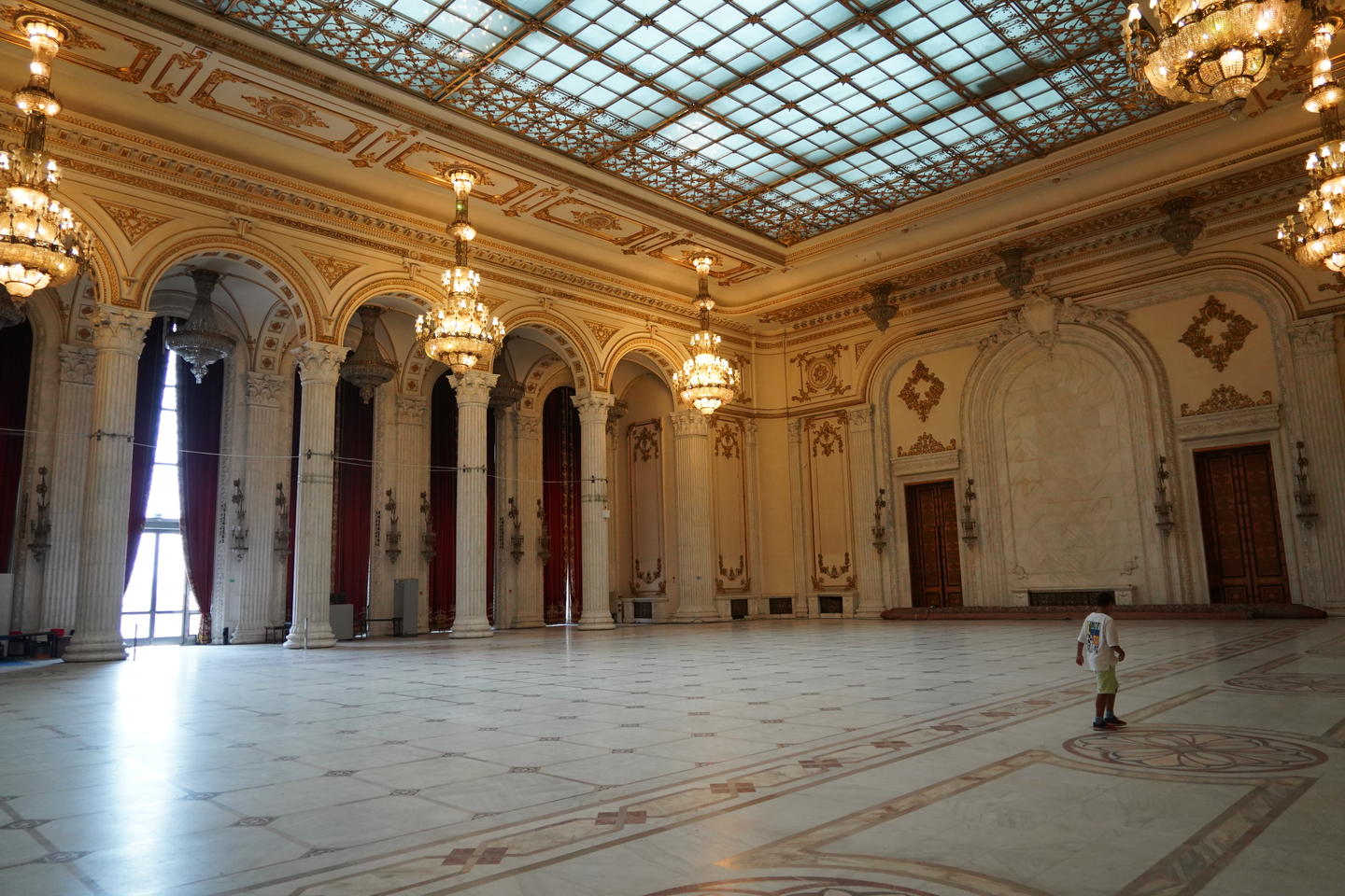 Interior view of Bucharest Palace of the Parliament with a steel-girded glass ceiling upheld by golden and marble columns, with tall red-draped windows and ornate crystal chandeliers.