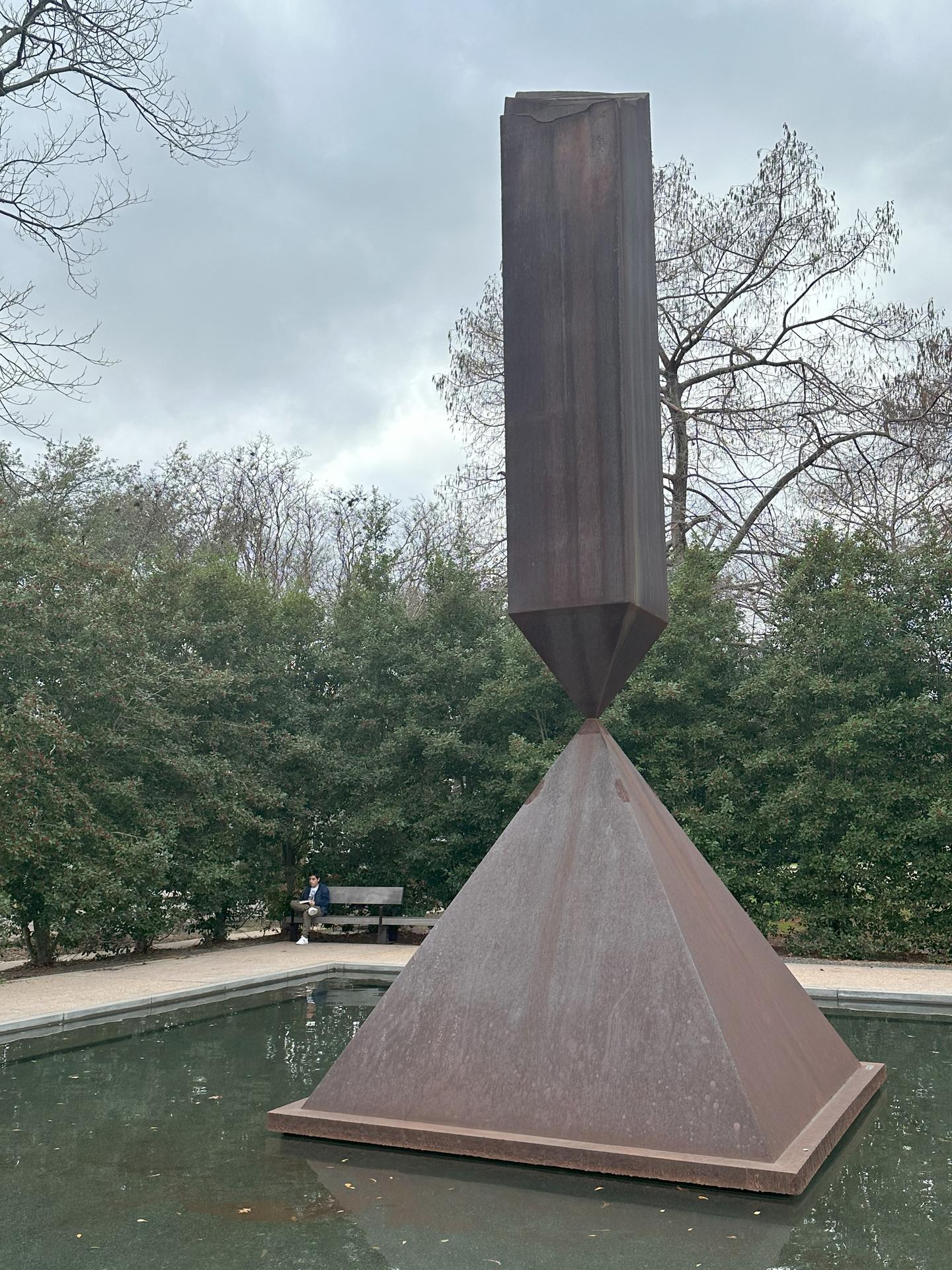 Rusted metal sculpture entitled "The Broken Obelisk" set in a shallow relfecting pond and surrounded by a square of tall evergreens in Houston
