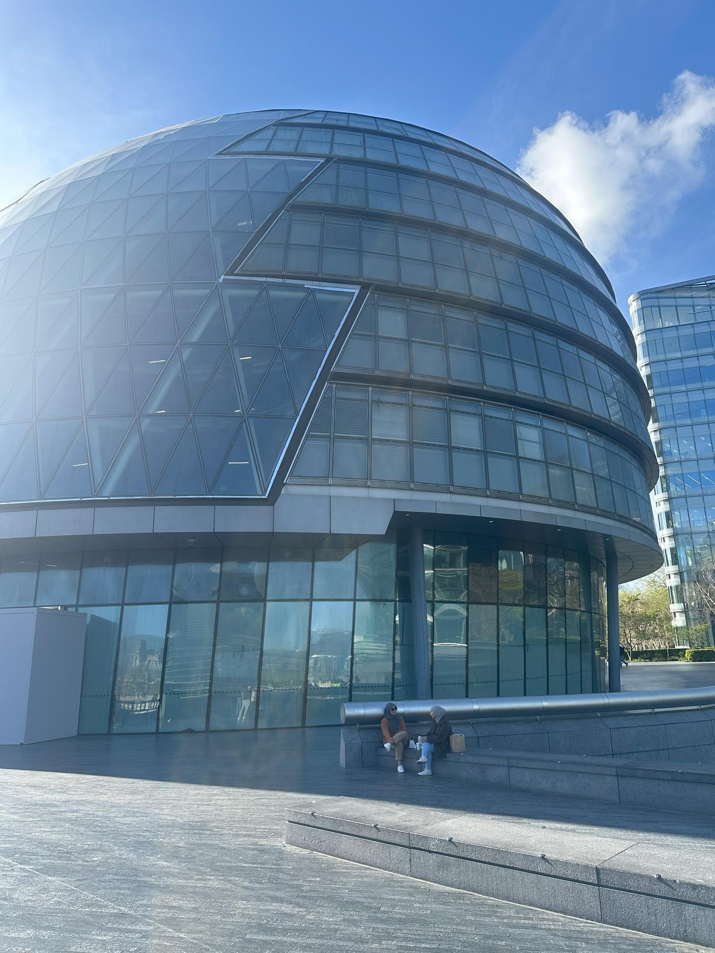 The glass curved dome of the office building of London City Hall sits under a blue sky.